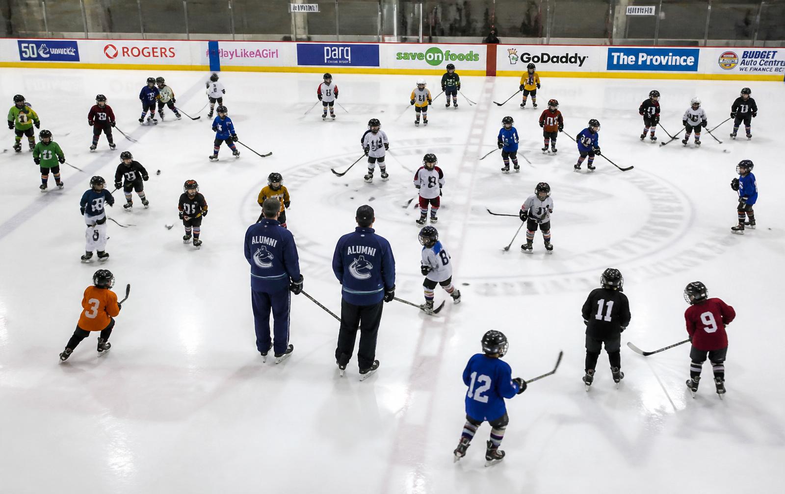 Overhead view of a large youth hockey clinic with dozens of kids and two Canucks Alumni coaches at center ice.