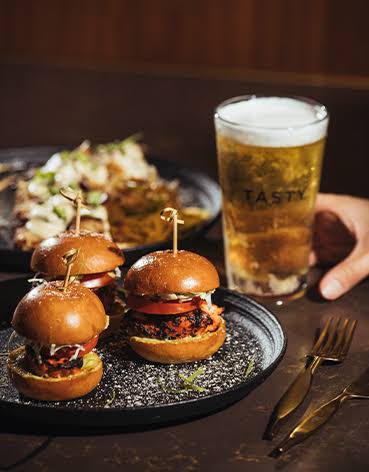 Trio of tandoori sliders on brioche buns served on a dark plate beside a pint of beer in a Tasty-branded glass.