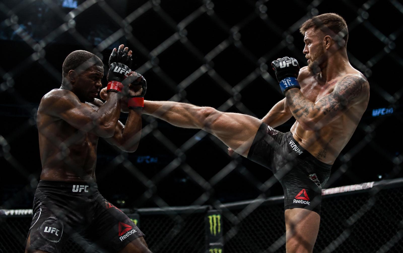 Two MMA fighters mid-exchange inside the octagon, one landing a head kick through the cage fence.